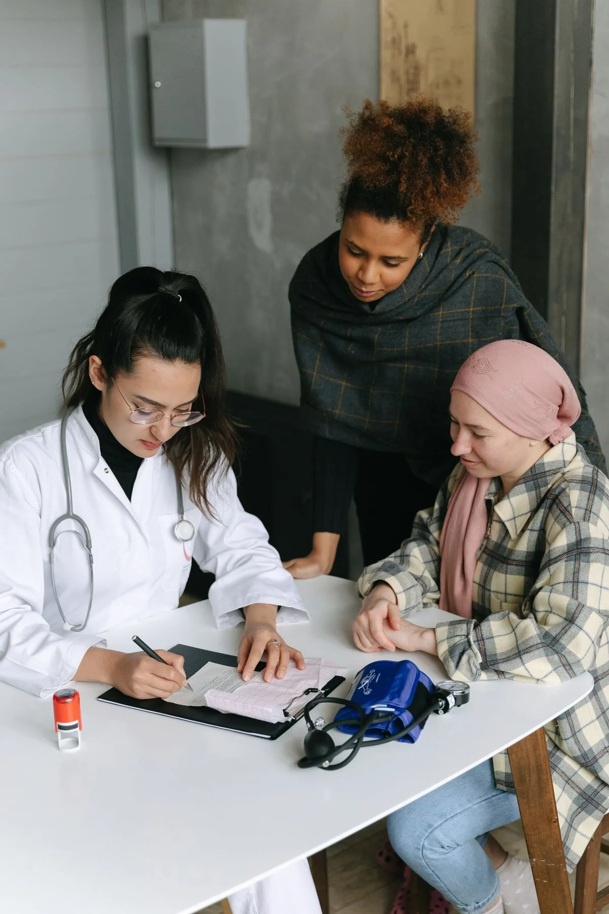 A person wearing a white coat and stethoscope writes at a desk while two people look on